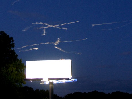 Dezorientovaní ptáci zmateně kroužící nad zdola nasvíceným billboardem